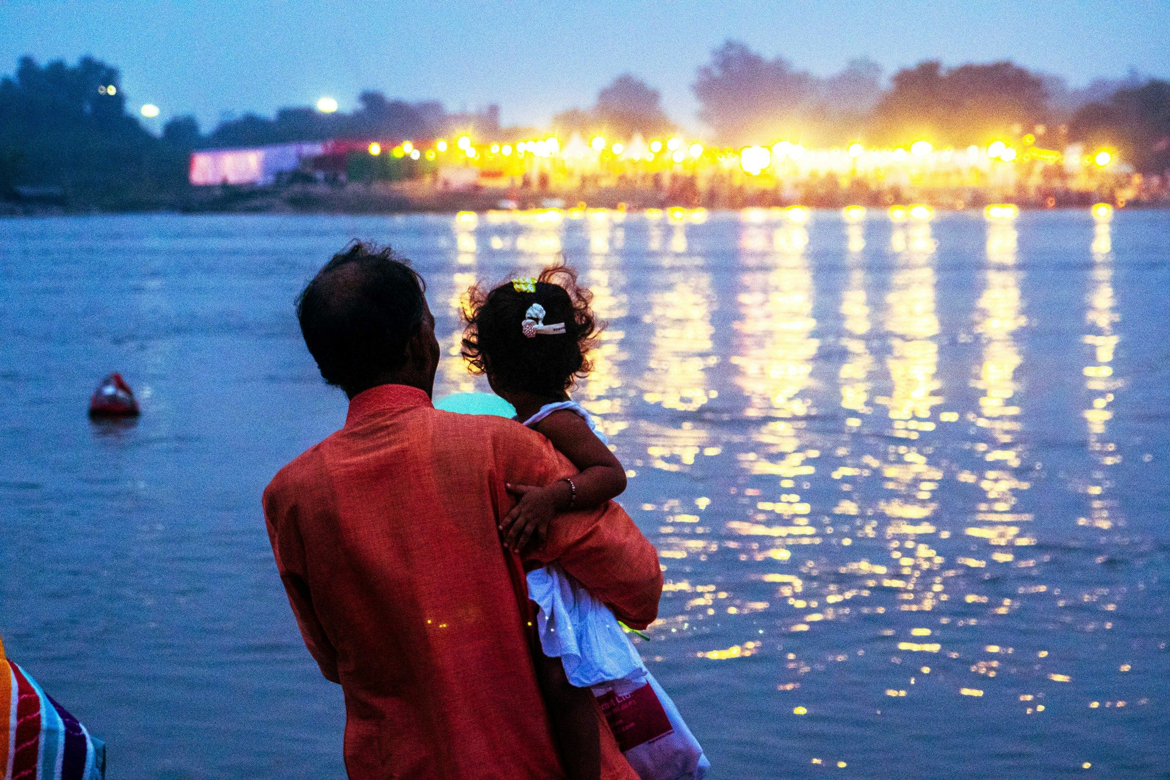 A father holding his young daughter by a river at dusk, city lights in the distance